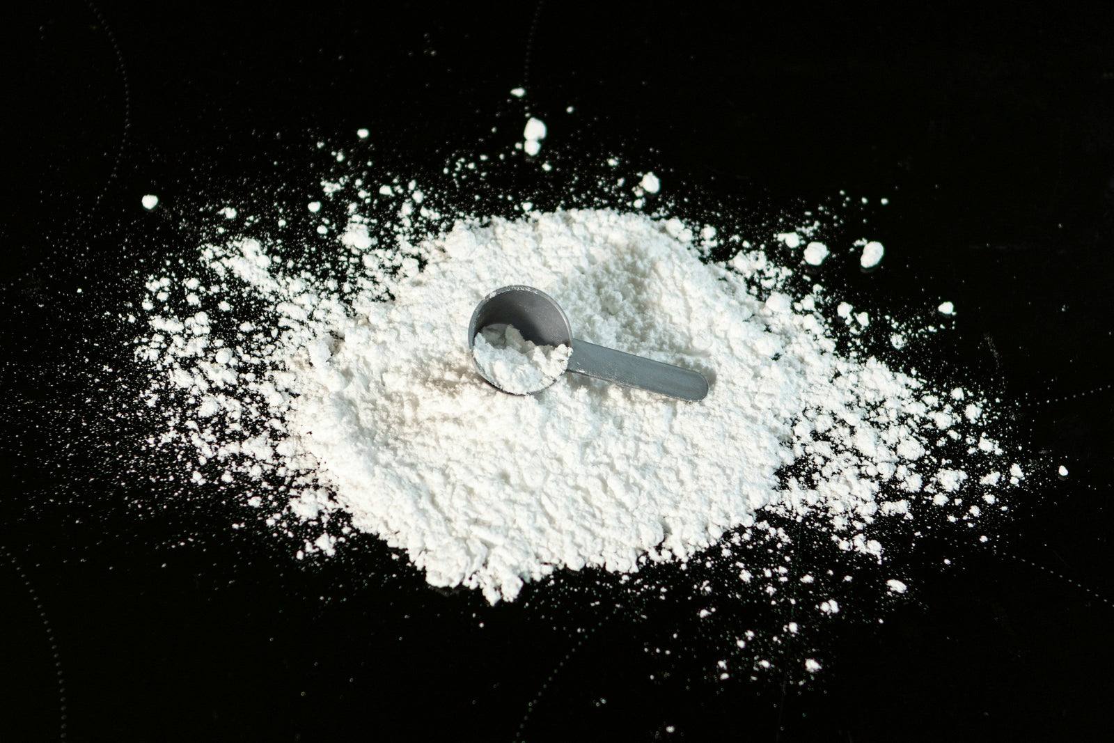 A small, metallic measuring scoop rests in a pile of white protein powder on a dark, possibly black, surface. Some of the protein powder is scattered around the main pile.